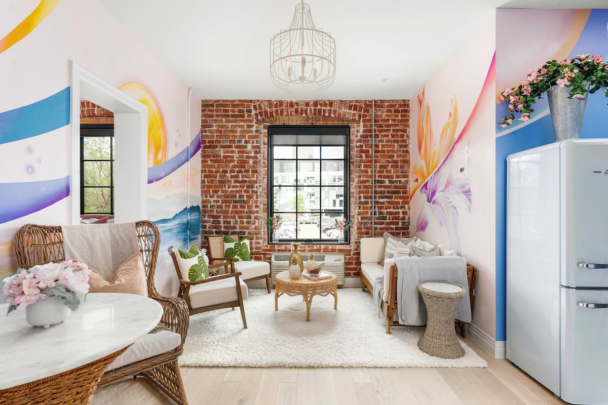 A cozy living room with a brick wall, a large window, wicker furniture, and a white refrigerator.