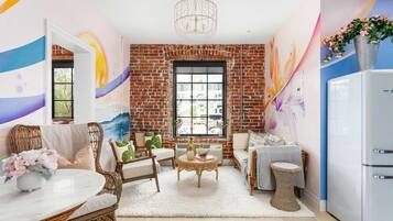 Una sala de estar acogedora con pared de ladrillo, un ventanal grande, muebles de mimbre y un refrigerador blanco.