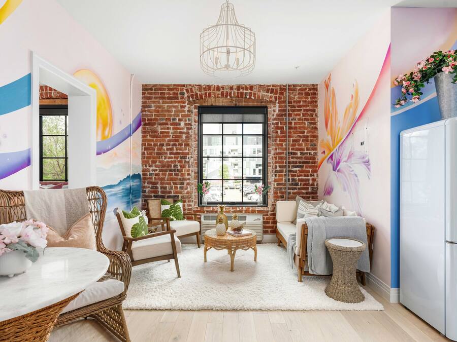 Una sala de estar acogedora con pared de ladrillo, un ventanal grande, muebles de mimbre y un refrigerador blanco.