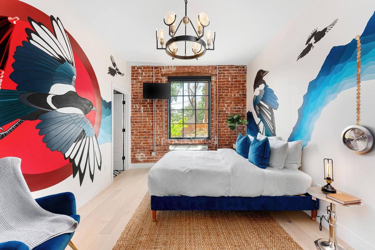 A bedroom with a brick wall, a large mural, a bed, a TV, and a chandelier.