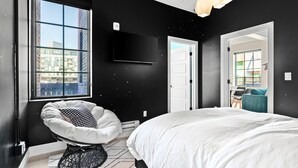 Ein modernes Schlafzimmer mit einem großen Bett, einem Sessel, einem Fernseher und Blick auf die Stadt durch ein Fenster.