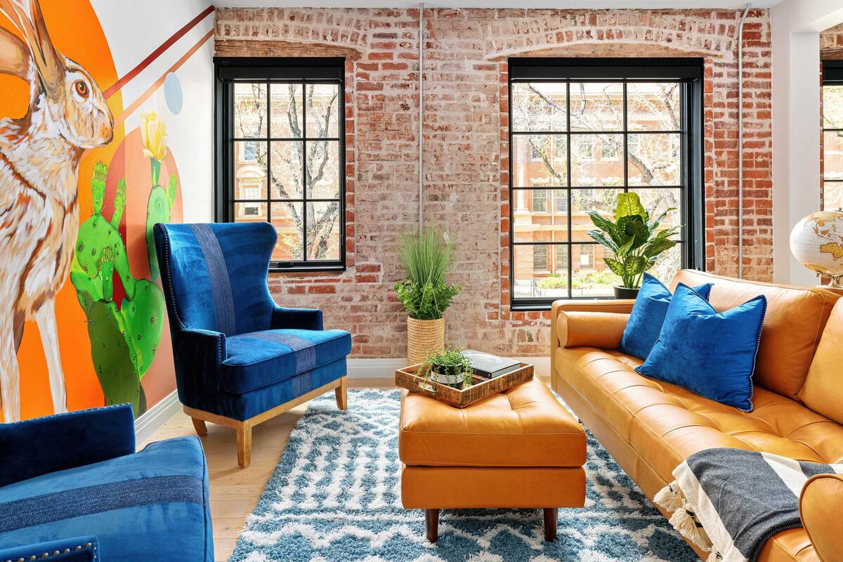 A living room with a brick wall, two windows, a blue armchair, an orange sofa, and a large cactus painting.