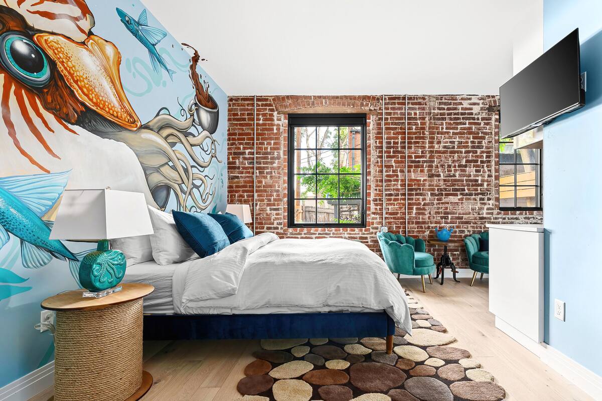 A modern bedroom with a brick wall, a large bed, teal chairs, and a mural of fish.