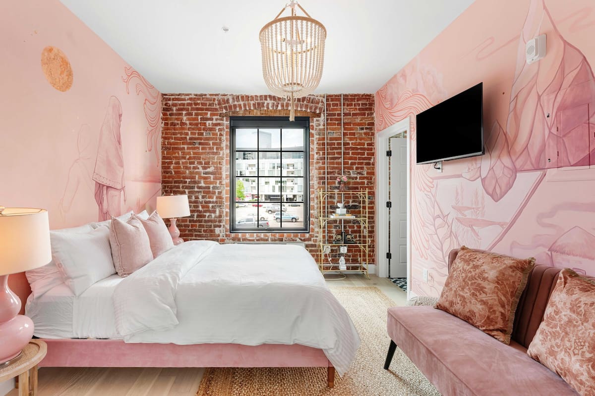 A bedroom with a brick wall, a large window, a bed, a TV, and a pink sofa.