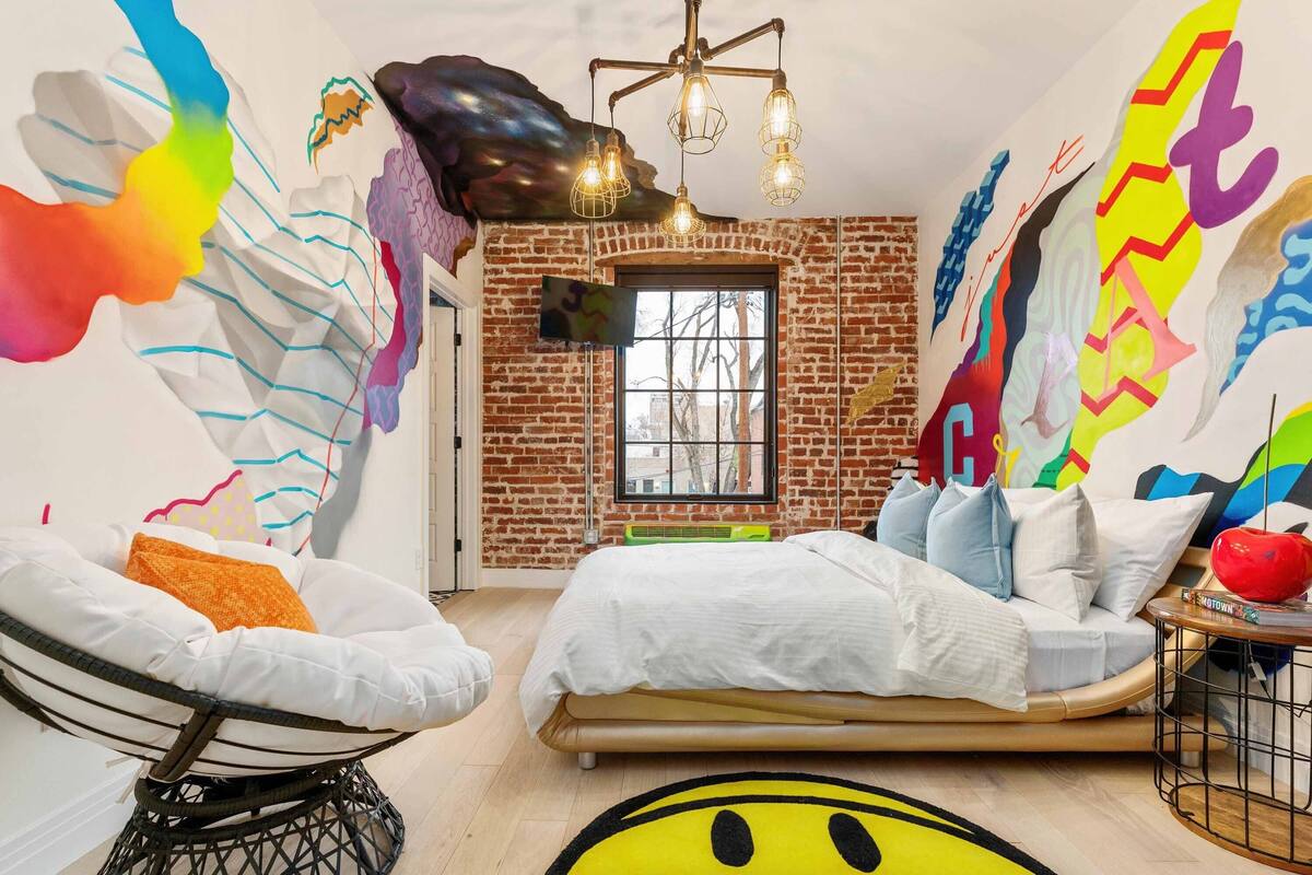 A bedroom with a brick wall, a bed, a chair, and a colorful abstract mural.