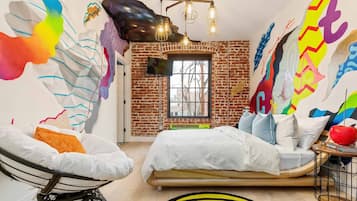 A bedroom with a brick wall, a bed, a chair, and a colorful abstract mural.