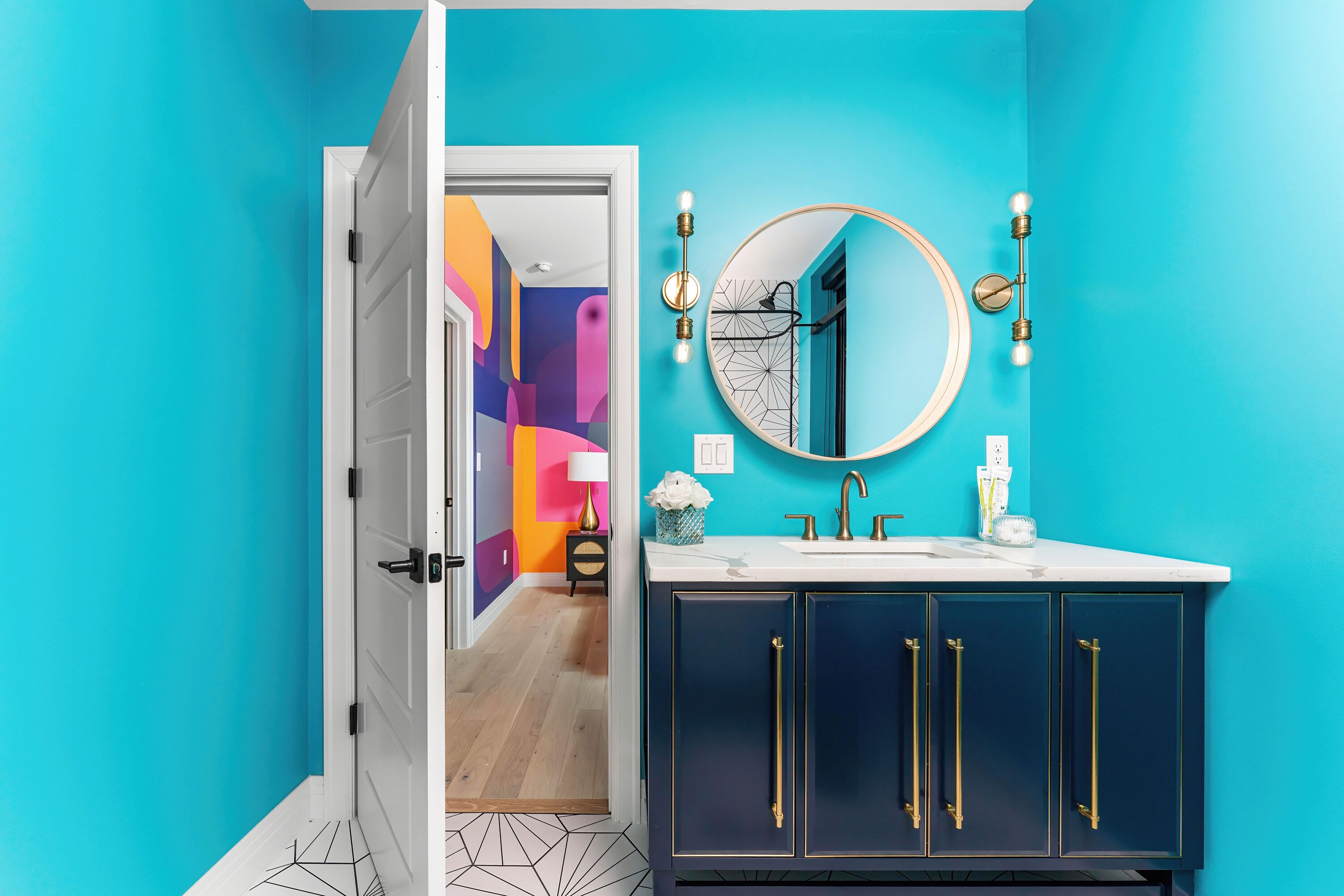 Une salle de bain aux murs de couleur turquoise, avec une porte blanche, un miroir rond et des meubles sombres dotés de poignées dorées.