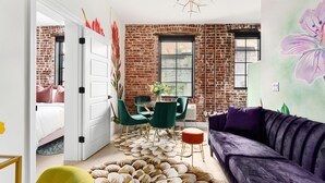 Ein modernes Wohnzimmer mit einer Ziegelwand, einem violetten Sofa, grünen Sesseln und einer weißen Tür, die zum Schlafzimmer führt.