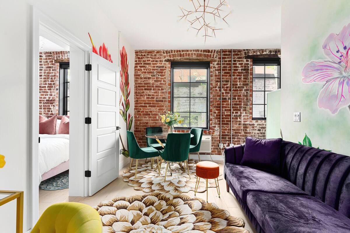 A modern living room with a brick wall, a purple sofa, green chairs, and a white door leading to a bedroom.