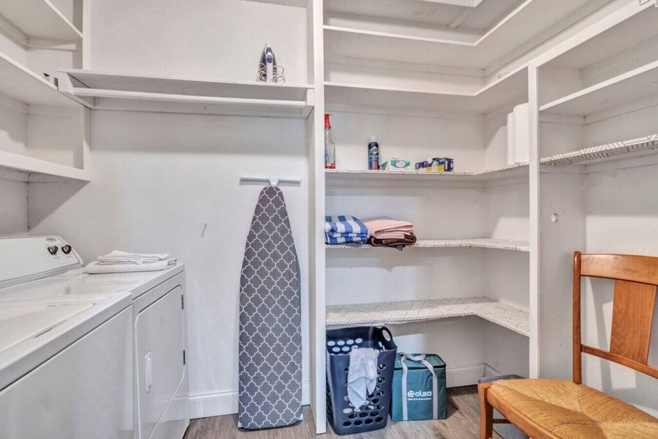 Un área de lavandería con lavadora, tabla de planchar y estantes con ropa doblada y productos de limpieza.