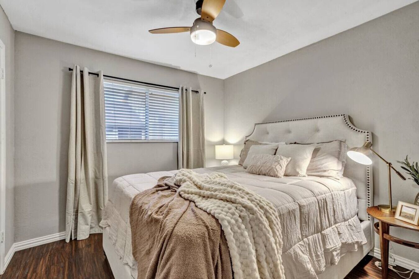 Une chambre à coucher avec un lit, un ventilateur de plafond, une fenêtre avec des rideaux et une table de chevet avec une lampe.