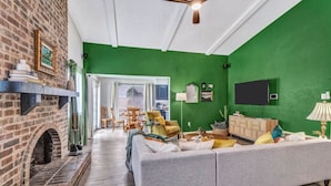 A living room with a brick fireplace, green walls, a ceiling fan, a sofa, and a TV mounted on the wall. - Pool Playhouse: FirePit, Grill, Billiards & Jenga (Richardson)