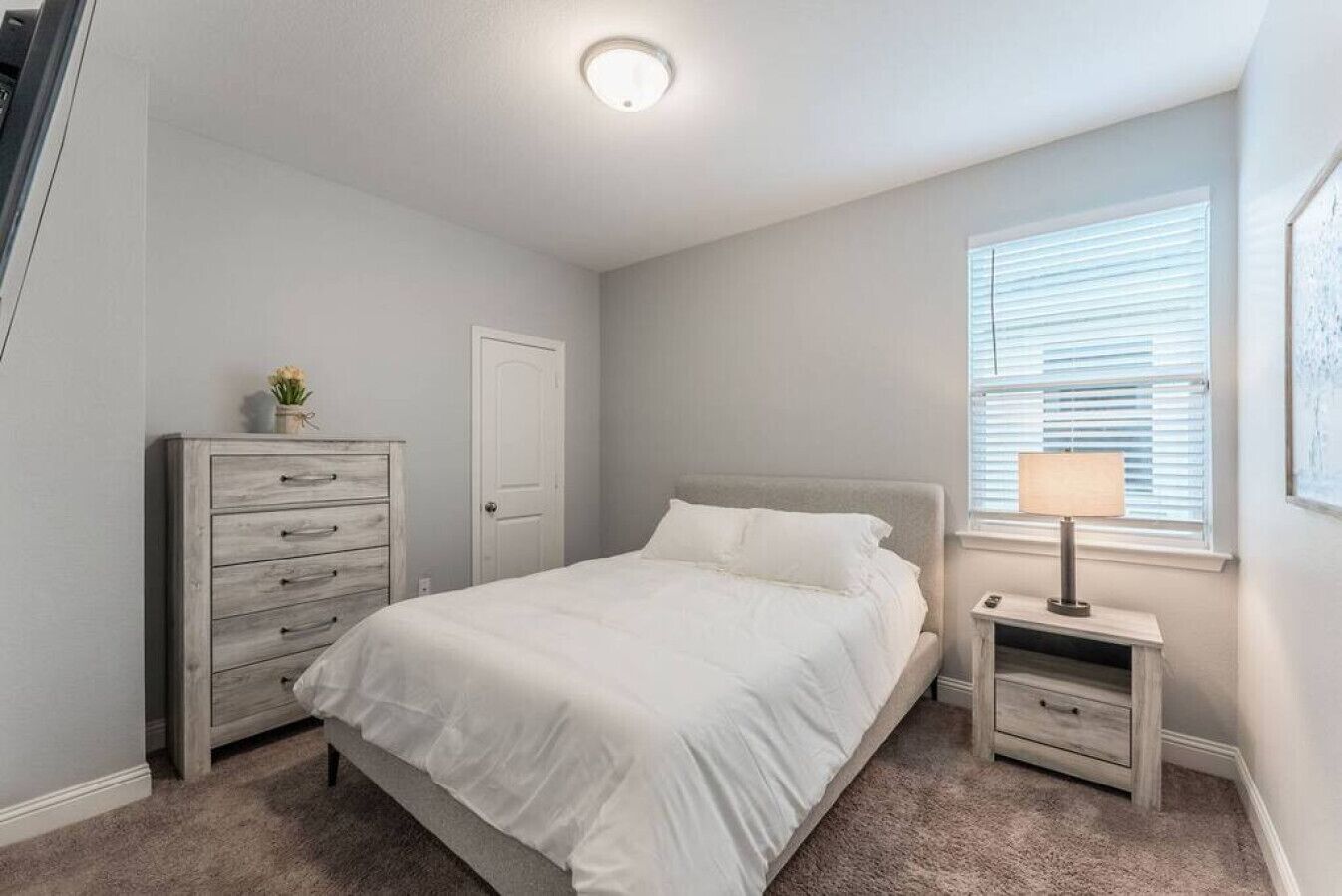 A bedroom with a bed, a dresser, a nightstand, a lamp, and a window with blinds.