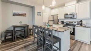 Uma cozinha moderna com armários brancos, uma ilha central com banquetas, uma área de jantar com mesa e cadeiras, e um lustre pendurado.
