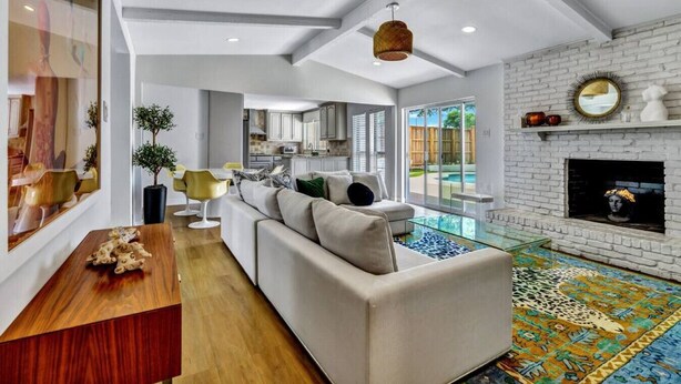Un salon moderne avec un canapé, une table basse et une cheminée. On aperçoit une cuisine au fond et, à l’extérieur, une piscine.