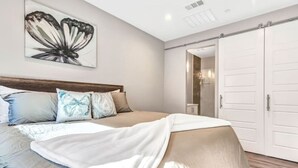 Ein Schlafzimmer mit Bett, Kissen, einem an der Wand hängenden Kunstwerk, einem Kleiderschrank und einer Tür zu einem anderen Raum.