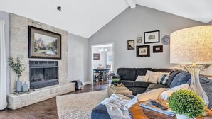 A living room with a stone fireplace, a dark-colored sofa, and a dining area in the background. - Pool PlayHouse: Firepit, Grill, Games & Sunlounger (Allen)