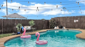 A backyard with a swimming pool, two pink flamingo floats, a striped umbrella, and a wooden fence. - Pool PlayHouse: Firepit, Grill, Games & Sunlounger (Allen)