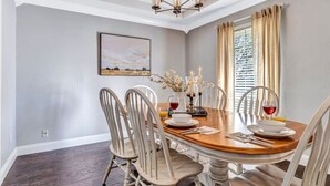 A dining room with a round table, white chairs, a chandelier, and a window with curtains. - Pool PlayHouse: Firepit, Grill, Games & Sunlounger (Allen)