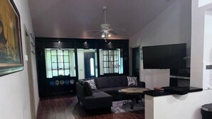 Una sala de estar con un sofá oscuro, una pequeña mesa de centro y un televisor montado en la pared. Hay un ventilador de techo sobre el sofá y una ventana con persianas al fondo.