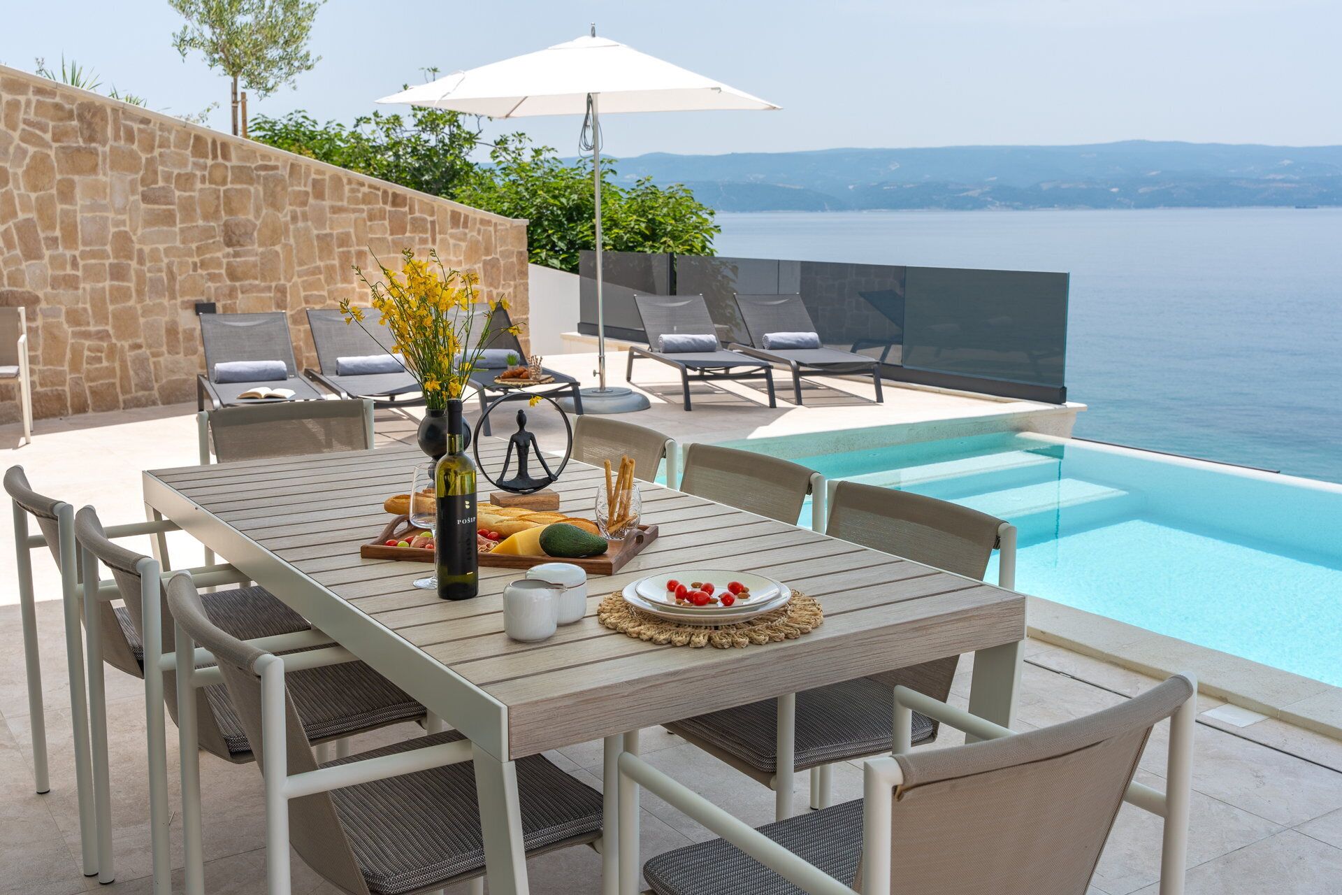 A poolside dining area with a table set for two, a bottle of wine, and a vase of flowers.