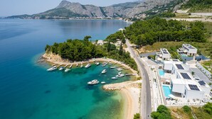 Una zona costera con barcos amarrados en un puerto deportivo, una carretera que conduce a un edificio moderno con paneles solares y un telón de fondo montañoso.