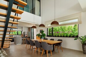 Un espace de repas moderne avec une table et des chaises en bois, une cuisine en arrière-plan et de grandes fenêtres donnant sur un espace verdoyant.