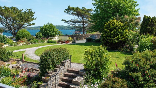 Een tuin met een stenen trap naar een zithoek, omgeven door weelderige begroeiing en uitzicht op zee.