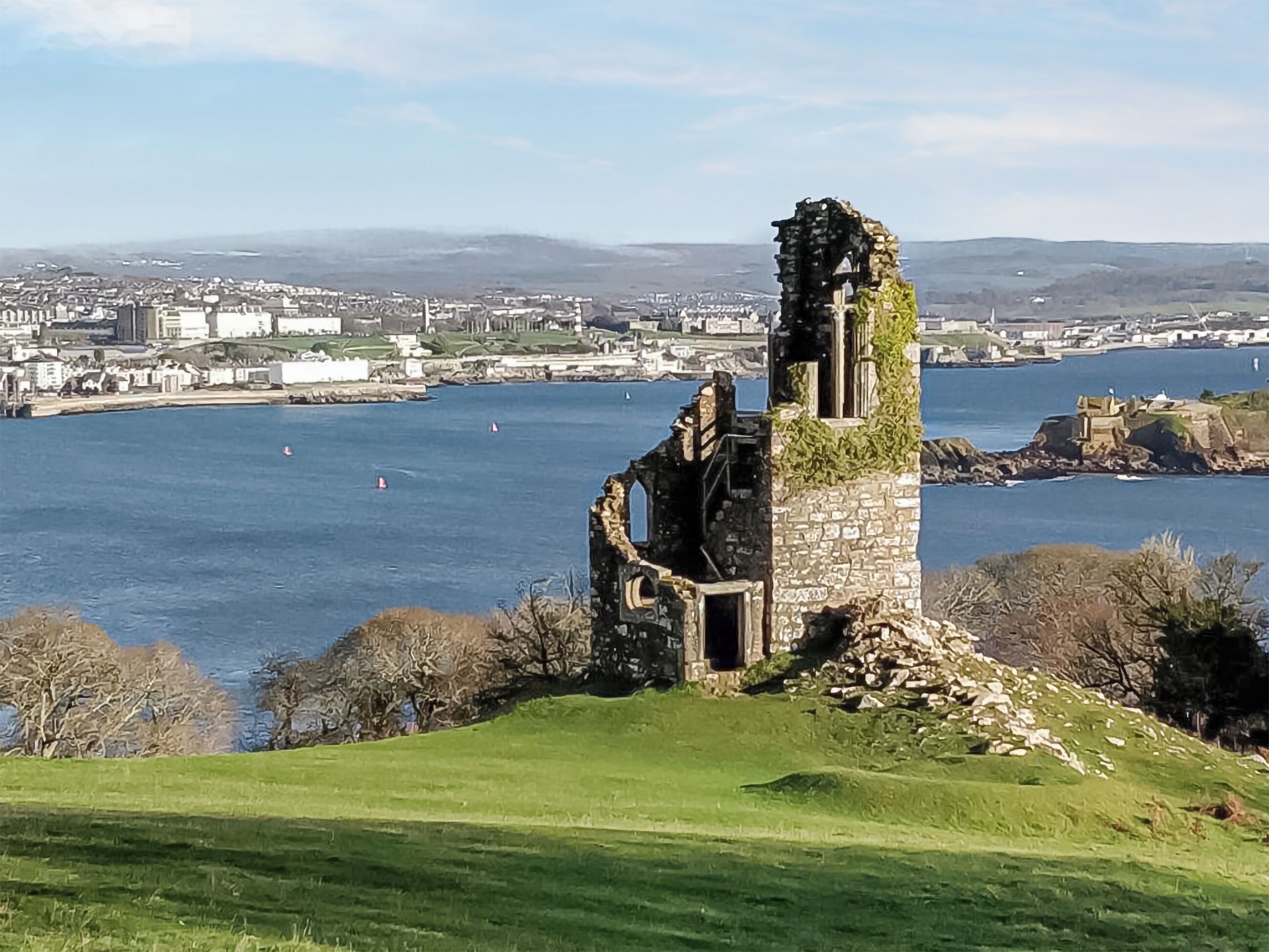 Ein historischer Stein-Turm mit einer Ruine, der auf eine Küstenstadt und ein Gewässer blickt.