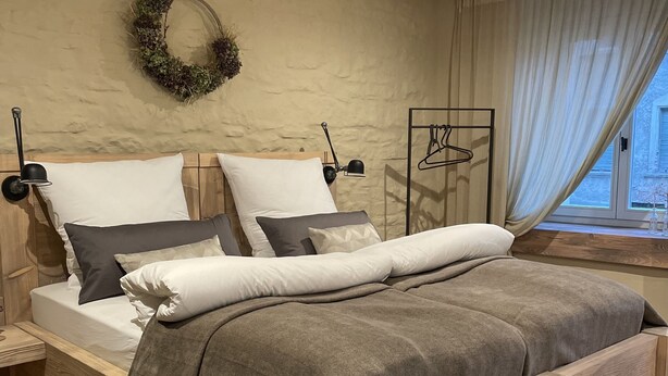 A neatly arranged bedroom with a wooden bed, white and gray bedding, a wall-mounted light, and a window with sheer curtains. - Unterkunft Nähe Magdeburg und Oschersleben Motorpark (Wanzleben-Börde)