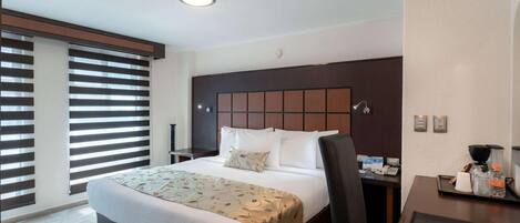 A hotel room with a large bed, a desk with a chair, a window with blinds, and a wall-mounted phone.