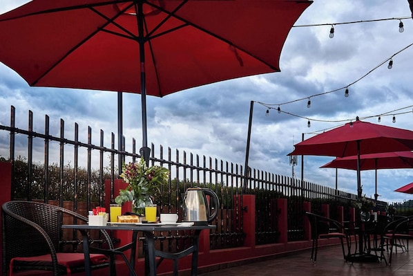 An outdoor dining area with red umbrellas, a table with breakfast items, and a metal fence in the background. - Sleep Inn Puebla Centro Historico (Puebla)