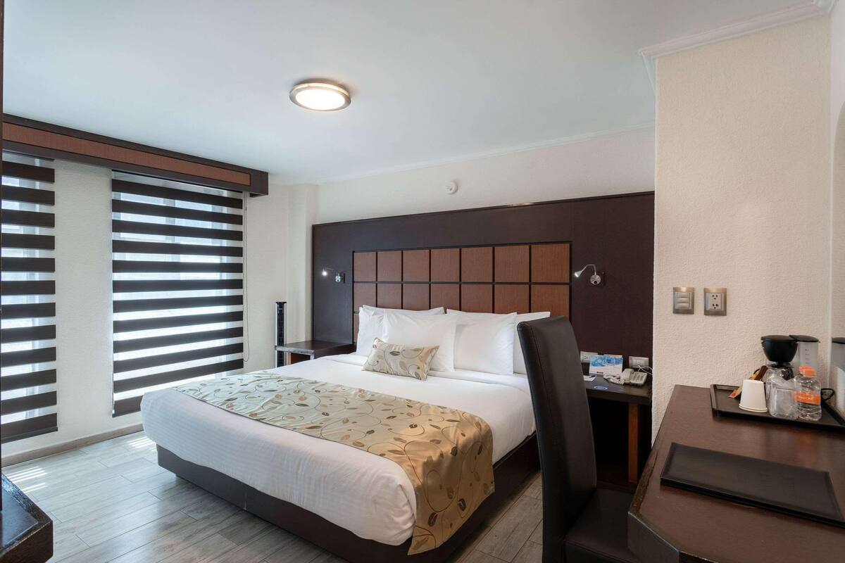 A hotel room with a large bed, a desk with a chair, a window with blinds, and a wall-mounted phone.