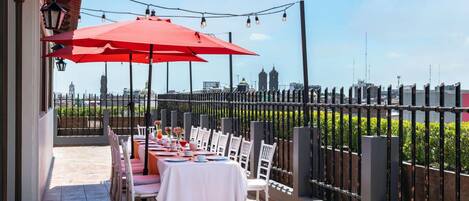 Un área de comedor al aire libre con mesas y sillas, un paraguas rojo y vista a la ciudad.