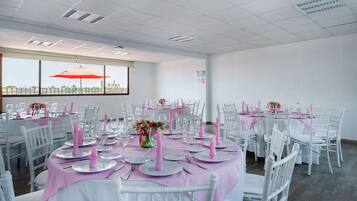 Een feestzaal met ronde eettafels, witte stoelen en een groot raam met een parasol.