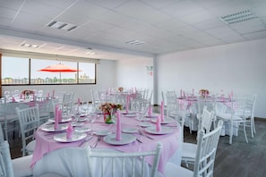 A banquet hall with round tables set for dining, white chairs, and a large window with an umbrella.