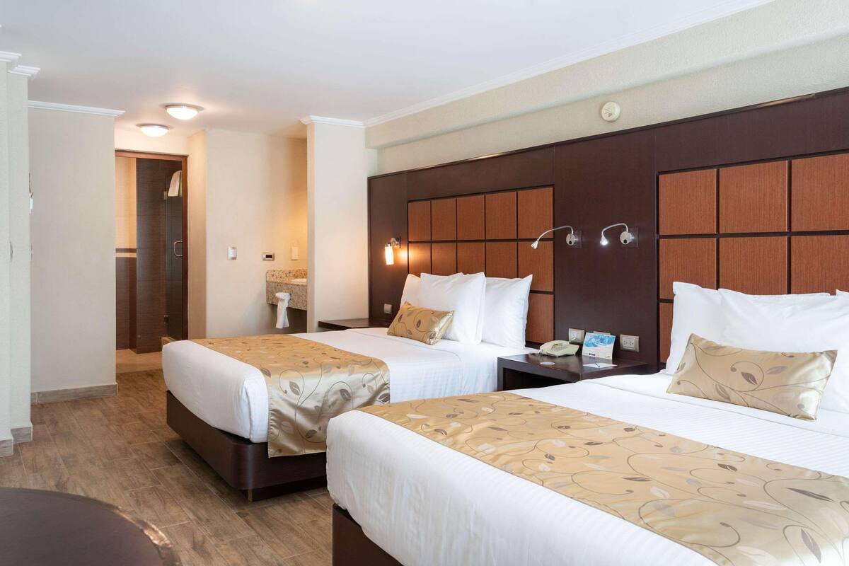 A hotel room with two beds, a wooden headboard, bedside lamps, and a nightstand with a phone and a magazine.
