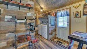 A rustic kitchen with wooden walls, a coffee station, a washing machine, and a window with curtains.