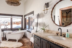 Een badkamer met een vrijstaand bad, een wastafel met een marmeren blad, een grote spiegel en uitzicht op een besneeerd landschap door een raam.