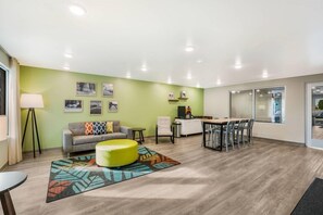 A modern living room with a sofa, ottoman, and a dining area with chairs and a table. - Woodspring Suites Downers Grove - Chicago (Downers Grove)