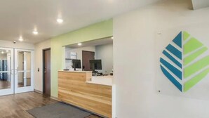 A reception area with a wooden reception desk, a glass door entrance, and a logo on the wall. - Woodspring Suites Downers Grove - Chicago (Downers Grove)