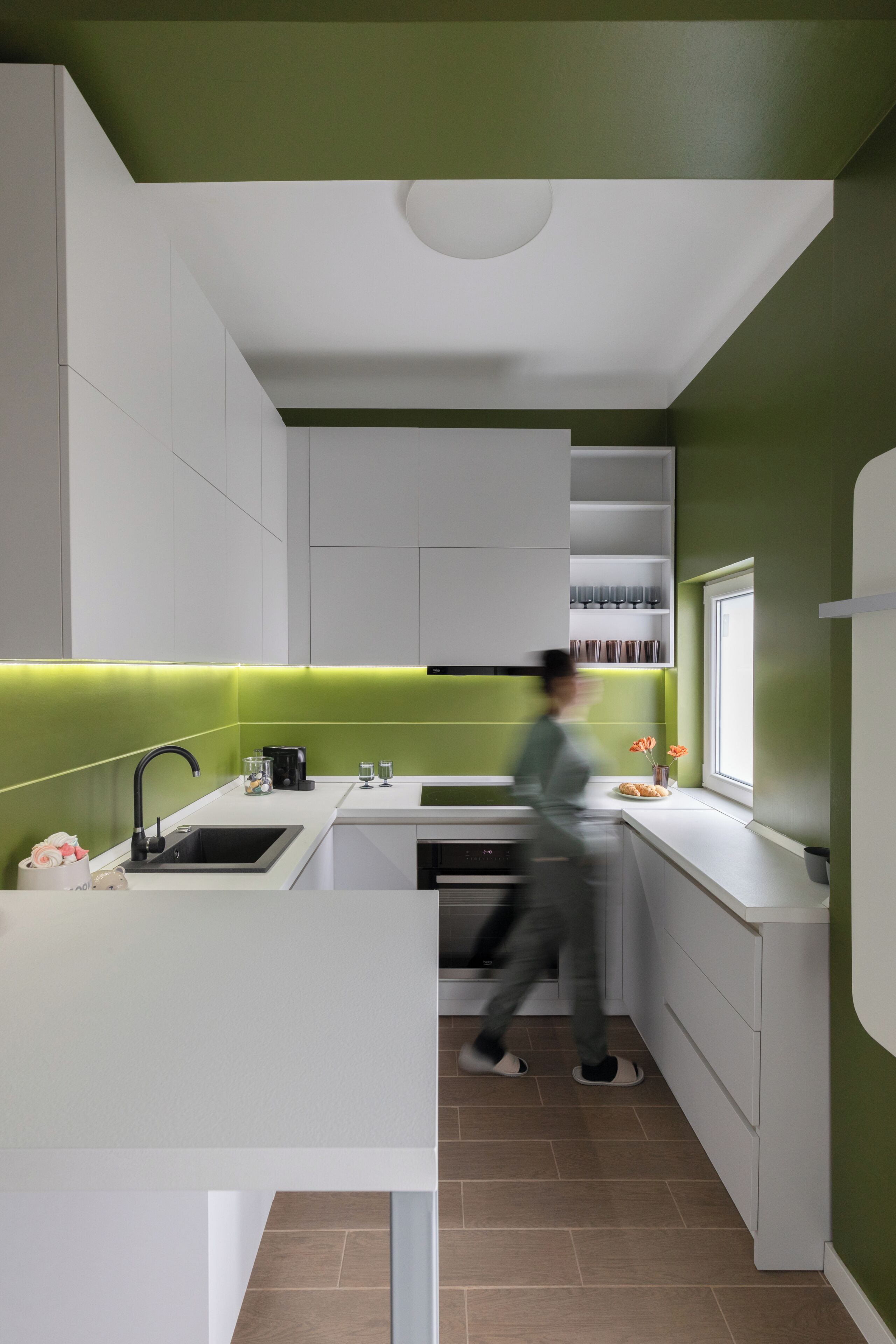 A modern kitchen with white cabinetry, a green accent wall, a window, and a person walking.