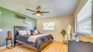 Une chambre à coucher comprenant un lit, un ventilateur de plafond, une fenêtre avec des stores, une table de chevet et un miroir fixé au mur.