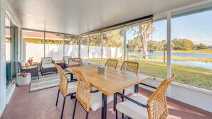Une terrasse couverte avec une table en bois et des chaises en osier, donnant sur une étendue d’eau et des arbres.