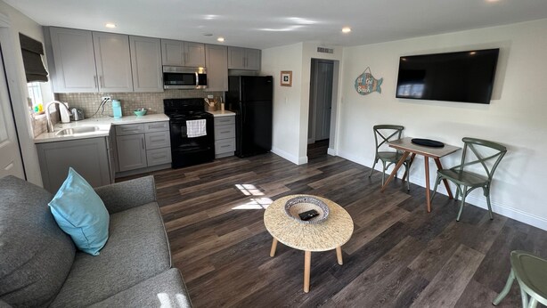A modern kitchen with grey cabinets, a black refrigerator, and a microwave. There is a dining area with a round table and two chairs. A flat-screen TV is mounted on the wall.