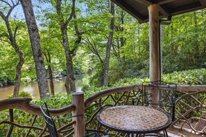 A wooden deck with a metal table and chair overlooking a river surrounded by lush greenery. - Highlands cabin on scenic pond, 6 min drive to Main Street & pet friendly! (Highlands)