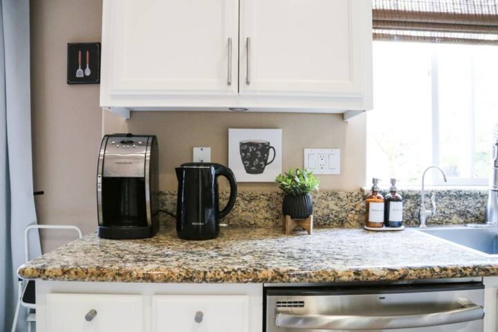 A kitchen with white cabinets, a coffee maker, a kettle, a sink, and a countertop with a plant and two bottles.