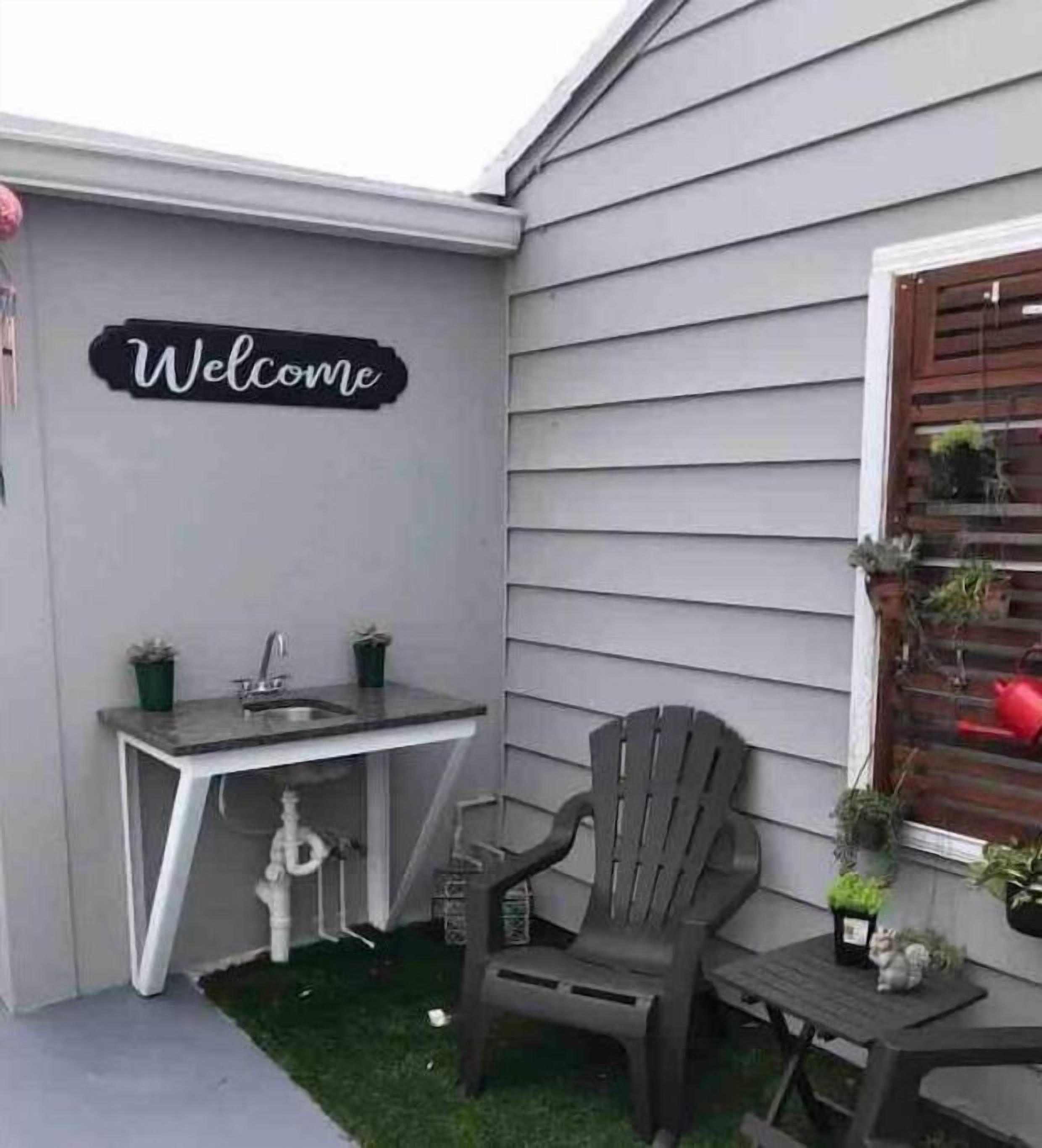 Eine kleine Terrasse im Freien mit einem Willkommensschild, einem Waschbecken, zwei Stühlen und Blumentöpfen.