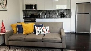 A modern living room with a grey sofa, a patterned throw pillow, and a red and white striped pillow. A kitchen with white cabinets, a stainless steel refrigerator, and a microwave is visible in the background. - Spacious Apartment Central Located (Hialeah)
