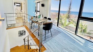 A modern interior with a dining area, staircase, and large windows offering a view of the sea and buildings. - Haus am Meer by Your Madeira Rentals (Paúl do Mar)